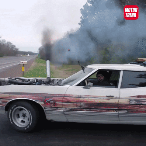 Video gif. Heavily modified car pulls onto and drifts onto a racetrack, black smoke pouring out of a tube linked directly into the exposed engine bay. We then see the car compete in a drag race against a similarly modified car.