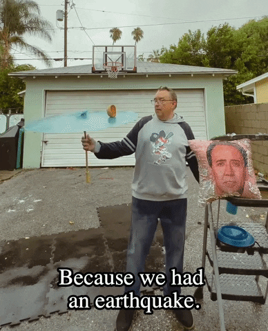 Video gif. Man stands in a driveway spinning a parasol as he lights a cheeseburger on fire that's spinning in place. He deftly keeps the cheeseburger in motion, looking dazzled as he talks. The man is standing next to a ladder that is randomly displaying a large shiny pillow with Nicholas Cage's face on it. Text, "Because we had an earthquake. 