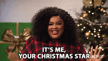 TV gif. Standing in front of a Christmas tree, Nicole Byer of Nailed It smiles flirtatiously and says, “It’s me, your Christmas star.”