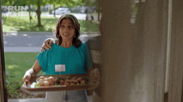 TV gif. The front door opens to an eager Andrew Phung as Andrew and Rakhee Morzaria as Camille in Run the Burbs who offer a tray of food and say together, “Welcome to the neighborhood!”