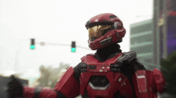 Video game gif. Red spartan from Halo stands on the side of a city street with his thumb out to hitchhike as cars drive past.