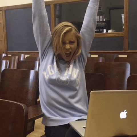 Studying -Video gif. A young woman sits in a classroom as she raises her hands in an excited cheer. 