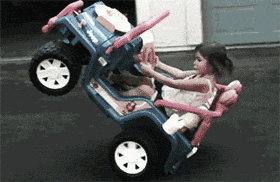 Video gif. A toddler sits in a ride on toy jeep, casually popping a wheelie in a driveway while she drives by. 