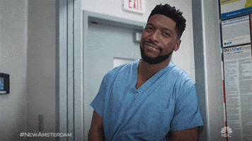TV gif. Jocko Sims as Dr. Floyd Reynolds in New Amsterdam leans in a doorway and gives us a double hello wave with his hands. He’s wearing hospital scrubs and has a face mask wrapped around his fingers.