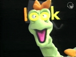 Video gif. A green dragon puppet with wide eyes and open mouth, and the word "look" is superimposed over his eyes like glasses.