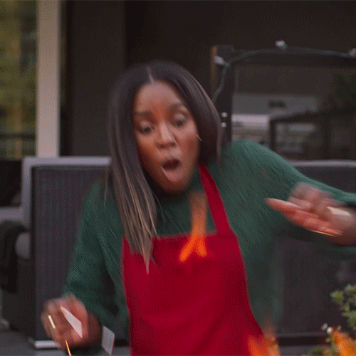 Movie gif. Wearing an apron, Kelly Rowland as Jacquie in Merry Liddle Christmas jumps back fearfully from a roaring fire.