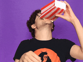 Video gif. A man tips his head and pours popcorn from a bag into his mouth though most seems to spill, bouncing off his face.