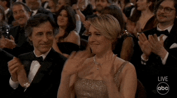 Oscars 2024 GIF. Greta Gerwig, seated at the Oscars and surrounded by applause, waves her hands girlishly in nervous gratitude, churching her hands to ground herself in the praise.