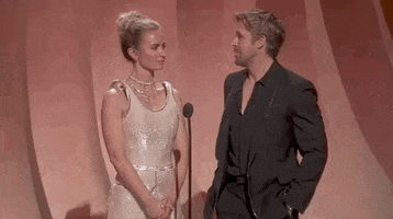 Oscars 2024 gif. Emily Blunt wears a pale gold dress with shoulder straps that hover over her shoulders, and Ryan Gosling wears a fully black tuxedo. Blunt stares at Gosling with a furrowed brow while Gosling peers at her out of the corner of his eye and cautiously says, "Hello Emily."
