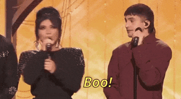 TV gif. Pentatonix on A Very Wicked Halloween stand on stage with their microphones. A woman yells, "Boo!" into her microphone towards a man who seems surprised even though he's looking directly at her. 