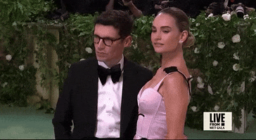 Met Gala 2024 gif. Closeup of Lily James posing for pictures. She dips a shoulder forward and back with a seductive expression. She's wearing a pale pink Erdem gown with a fitted bodice with a scalloped plunging neckline. A black vine with leaves detail weaves up and over her shoulders. 