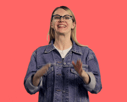 Video gif. In front of a solid coral background, a blonde woman in a jean jacket and glasses claps enthusiastically while grinning widely at us.