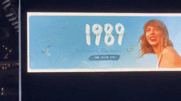 Video gif. Billboard ad for Taylor Swift's album 1989 Taylor's Version showing Taylor smiling and looking over her shoulder, wearing red lipstick as seagulls fly through a bright blue sky that fills the billboard.