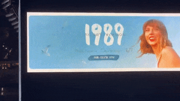 Video gif. Billboard ad for Taylor Swift's album 1989 Taylor's Version showing Taylor smiling and looking over her shoulder, wearing red lipstick as seagulls fly through a bright blue sky that fills the billboard.