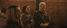 Movie gif. Olivia Colman as Hope and Jim Gaffigan as Zeke in "Them That Follow" sit amongst churchgoers; Hope raises her hand up with eyes closed and Zeke puts his hand on his knee and leans back.