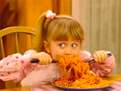 Animated GIF of a young toddler using a fork and spoon in each hand to fill her mouth with spaghetti.