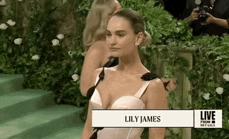 Met Gala 2024 gif. Closeup of Lily James posing for pictures with a serene expression. She's wearing a pale pink Erdem dress with a fitted bodice with a scalloped plunging neckline. A shimmery black vine with leaves weaves up through her bodice, peeking up over both shoulders from the back of the dress.