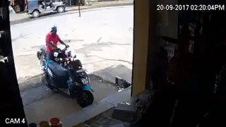 Video gif. A man on a motorcycle attempts to park his bike near a ledge, then tries to dismount but loses his balance. He falls over the ledge, his motorcycle falling on top of him. A crowd of people comes running to help him.
