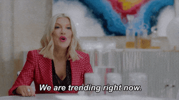 TV gif. Tori Spelling as herself from BH90210 wears a red jacket with a grid pattern. She speaks to us with a smile from across a table. Text, "We are trending right now."