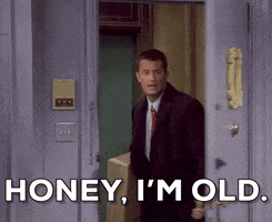 Friends gif. Matthew Perry as Chandler whips open the door to the apartment and says sarcastically, “Honey, I'm old.” He carries a large box in his hand. 