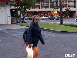 Video gif. A man stands in a parking lot wearing a coat and over ear headphone, holding a plastic shopping bag. He stops walking and smiles and gives an uncertain wave to someone off screen.