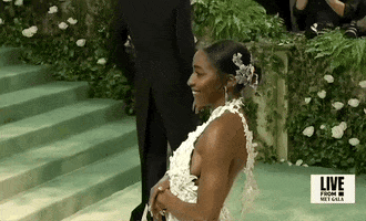 Met Gala 2024 gif. Ayo Edibiri wearing an unfussy Loewe gown of white 3D flower cutouts that fade into color, poses for the cameras, swinging one way, then the other, adjusting poses and offering cheeky looks.