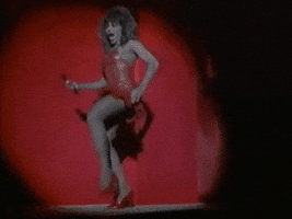 Video gif. Tina Turner does quick footwork across a stage, wearing a red mini dress and heels.