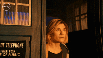 TV gif. Jodie Whittaker as Doctor Who stepping out of a phone booth into golden light, looking around.