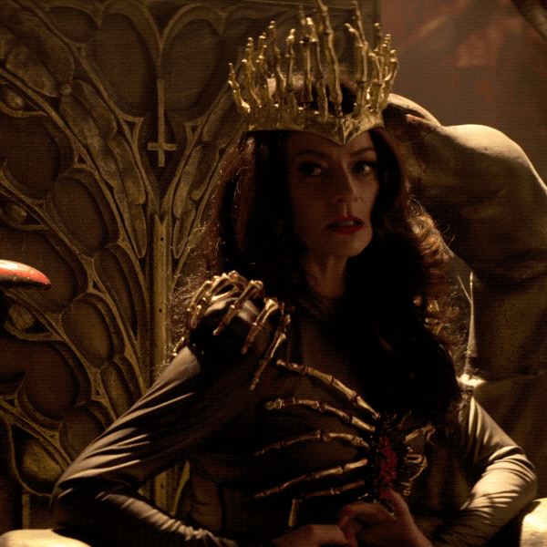 TV gif. Michelle Gomez as Madam Satan in Chilling Adventures of Sabrina tilts her crowned head curiously while twiddling her fingers, seated in an ornate golden throne