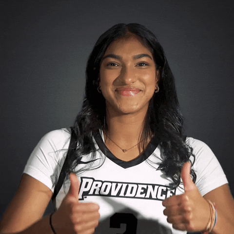 Video gif. Sahana Kanagasabay of the Providence College Women's Basketball team gives us two thumbs up and a big friendly smile against a dark gray background.
