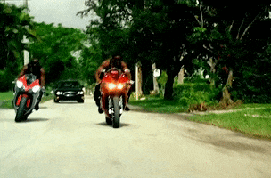 Music Video gif. Two muscular men in the Stuntin Like My Daddy music video ride on motorbikes as cars chase them down the road.