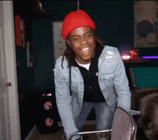 Video gif. Standing between a chair and a vertical metal pole in a darkly lit bedroom, a young man with dreadlocks in a red cap leans back for a laugh and falls gently onto his back. 