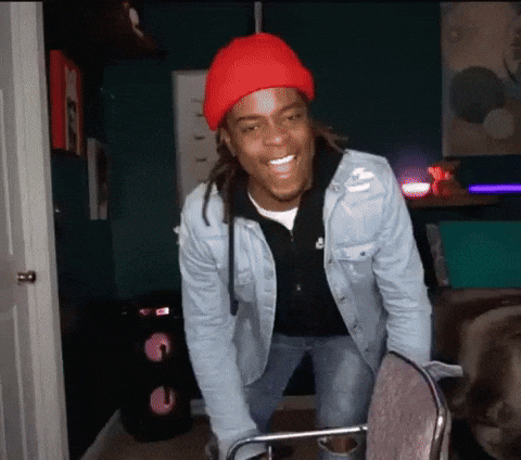 Video gif. Standing between a chair and a vertical metal pole in a darkly lit bedroom, a young man with dreadlocks in a red cap leans back for a laugh and falls gently onto his back. 