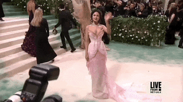 Met Gala 2024 gif. Eiza Gonzales wearing a quirky and ethereal Del Core gown, waves for the cameras with confidence.