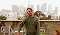 TV gif. Jake Johnson as Nick Miller in New Girl stands on a rooftop and victoriously pumps his fist in the air.