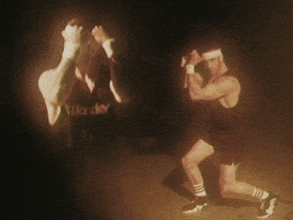 Video gif. Man is wearing workout clothes complete with sweatbands on his wrists and head. He puts his arms up in a fight position and does a couple lunges.