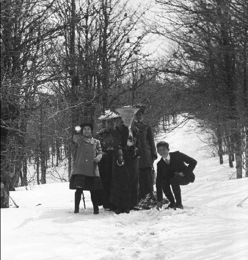 Video gif. Black and white vintage photo of a group of people standing in a snowy forest. With wide smiles, three people throw a snowball right at us, with the last one hitting us square in the face and morphing into text that reads, "Bonne année 2024!"