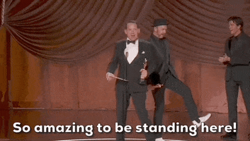 Oscars 2024 gif. Johnnie Burn gestures outwards with his arms as he holds the Oscars trophy and envelope. In disbelief, he says, "So amazing to be standing here!" Behind him, Tarn Willers wears a full black suit and points at his white shoes before pointing to someone in the crowd. 