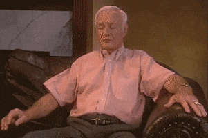 Video gif. An old man sits up straighter on a couch and waves his arms up and down like he is doing jumping jacks while seated.