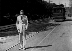 An animated gif that appears to be from a very old black and white movie with a man standing in the middle of a road. A streetcar on rails zooms towards him and at the last minute, turns instead of hitting him.