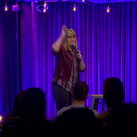 Celebrity gif. Christi Chiello stands on stage in front of a crowd of people. She looks a bit embarrassed and worried as she pulls the mic away from her mouth and raises her hand. 