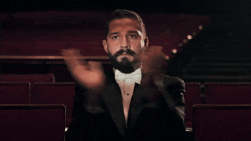 Meme gif. Shia LaBeouf sits in an empty auditorium in the audience. He has a serious expression on his face as he claps loudly.