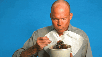 Video gif. A bald man wears a napkin tucked into his shirt and holds a bowl of food up to his chin as he takes a huge mouthful of stir fry vegetables and noodles, chewing and smiling.