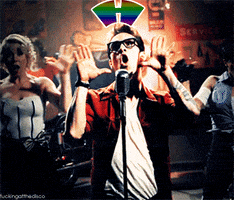Video gif. A man stands before a microphone, among other performers on stage, making a rainbow gesture with his hands as the word "homosexuality" appears in rainbow text.