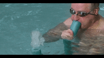 Music video gif. From Yung Bae's video for L.O.V.E., an old man wearing goggles in a swimming pool blows into a pool noodle, shooting water out the other end, which an older woman laughs at. The man smiles suggestively at us.