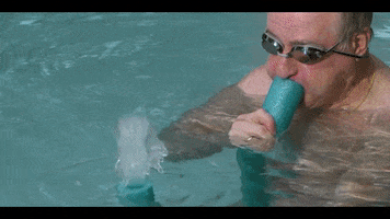 Music video gif. From Yung Bae's video for L.O.V.E., an old man wearing goggles in a swimming pool blows into a pool noodle, shooting water out the other end, which an older woman laughs at. The man smiles suggestively at us.