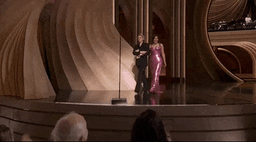Oscars 2024 GIF. Kate McKinnon and America Ferrera walk with linked arms towards the microphone. McKinnon is wearing an all black suit and Ferrera is wearing a Barbie pink sequined dress that fits her like a glove. 