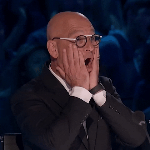 Reality TV gif. Howie Mandel from America's Got Talent holds his hands against the sides of his face as his mouth drops in shock. 