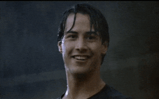 Movie gif. Keanu Reeves as Johnny Utah in Point Break smiles and gives a cool thumbs-up while standing soaking wet from the pouring rain around him.