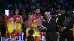 Sports gif. Jayson Tatum holds up the 2023 NBA All-Star Game MVP award and smiles as he takes a photo, the rest of his teammates watching as cameras flash.